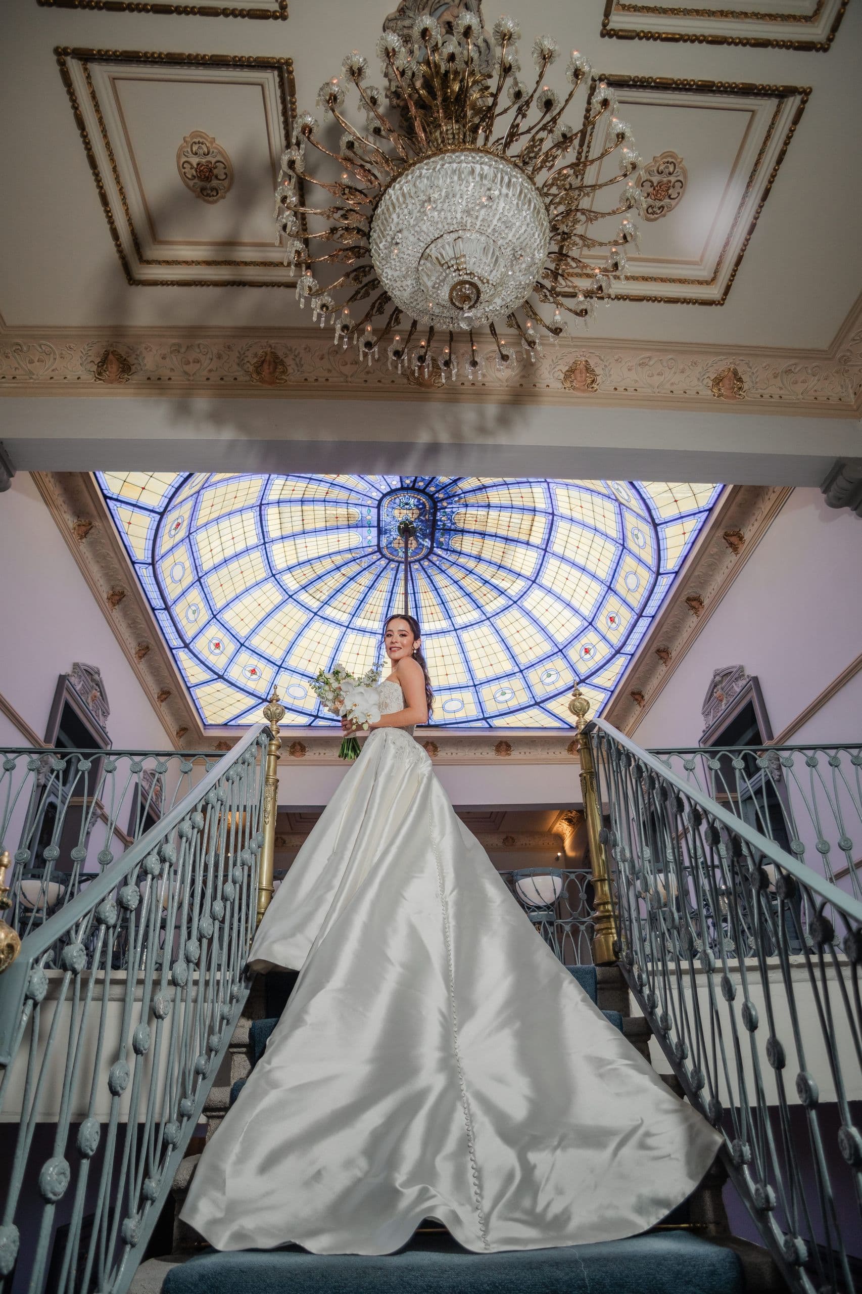 Bride with her bouquet on the staircase of Palacio Julio, beneath the leaded stained-glass dome