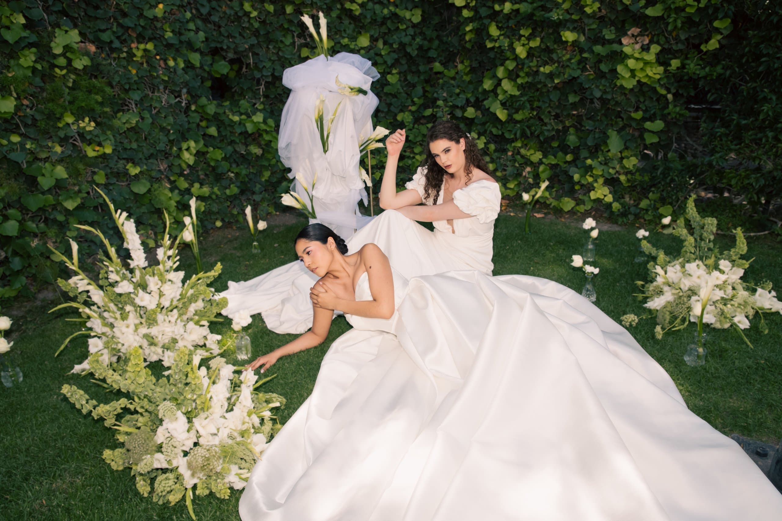 Dos novias sobre el pasto entre arreglos de gladiolas blancas y un muro de hiedra