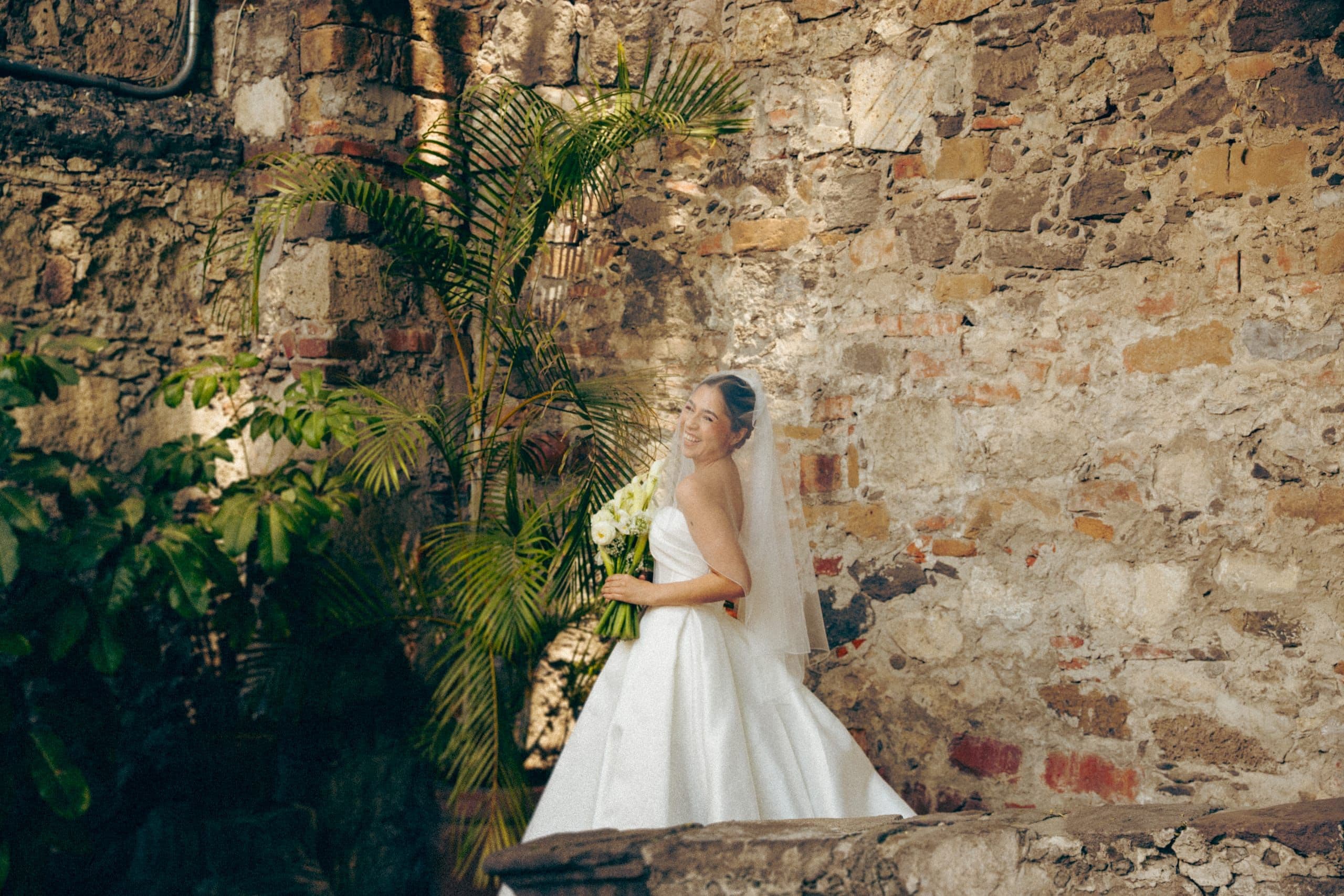 Novia recargada en un muro de piedra con helechos, el día de su boda en Puebla