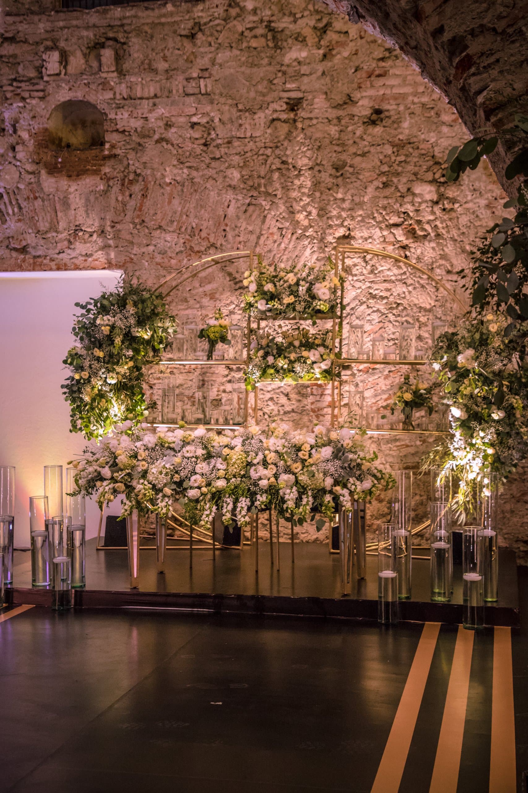 Muro de piedra iluminado con una instalación floral en tonos claros sobre la pista