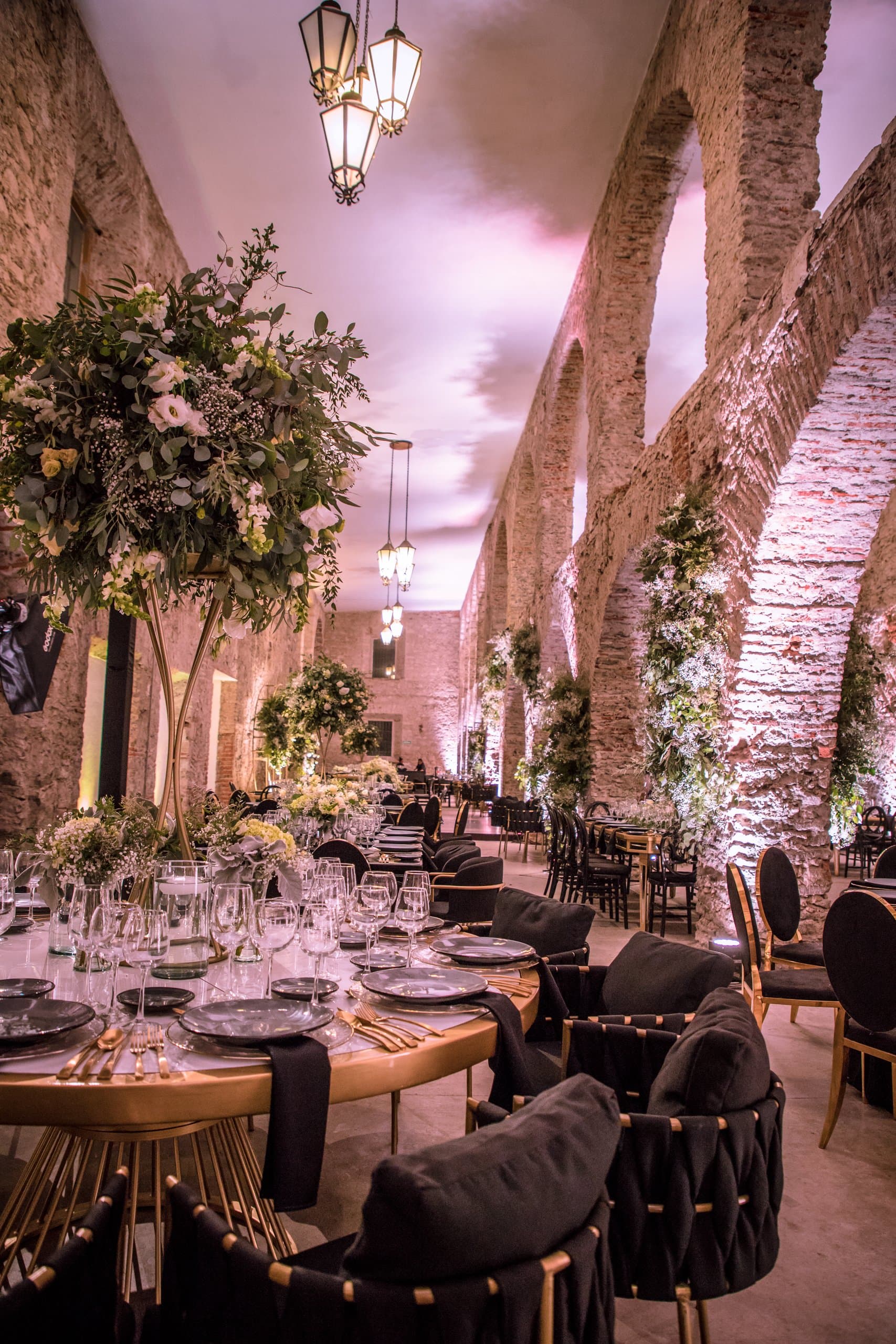 Salón bajo arcos de piedra con luz malva, árboles y mesas montadas por Clhei