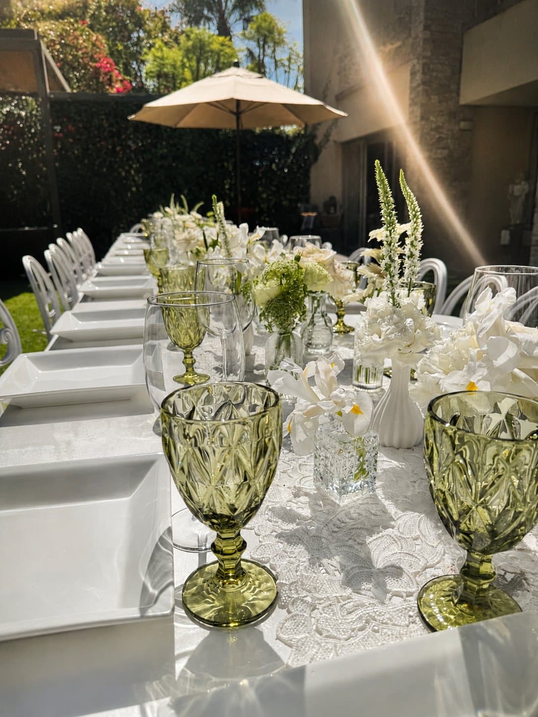 Mesa larga en blanco con cristalería verde y flor blanca, montaje de jardín