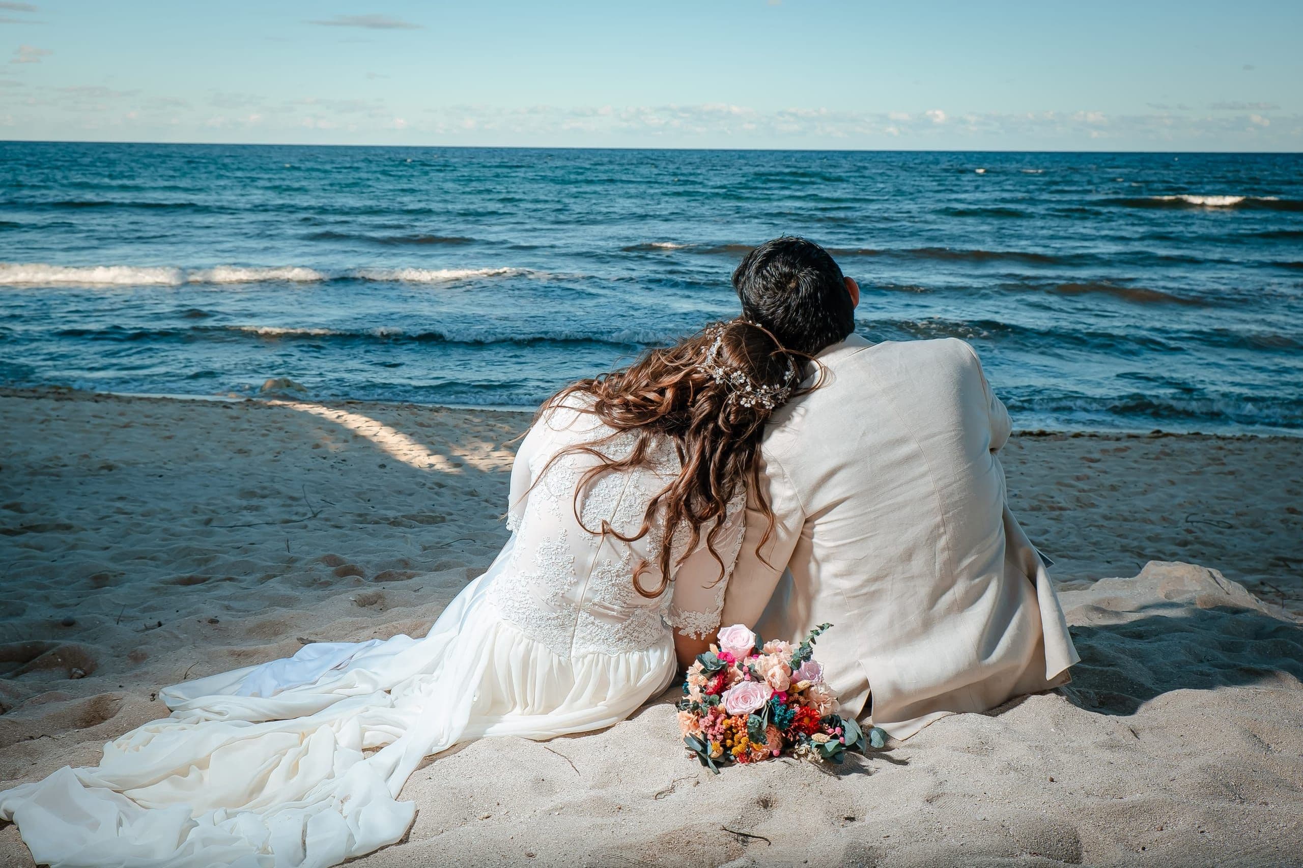 Pareja recién casada sentada en la playa frente al mar, con su ramo de colores