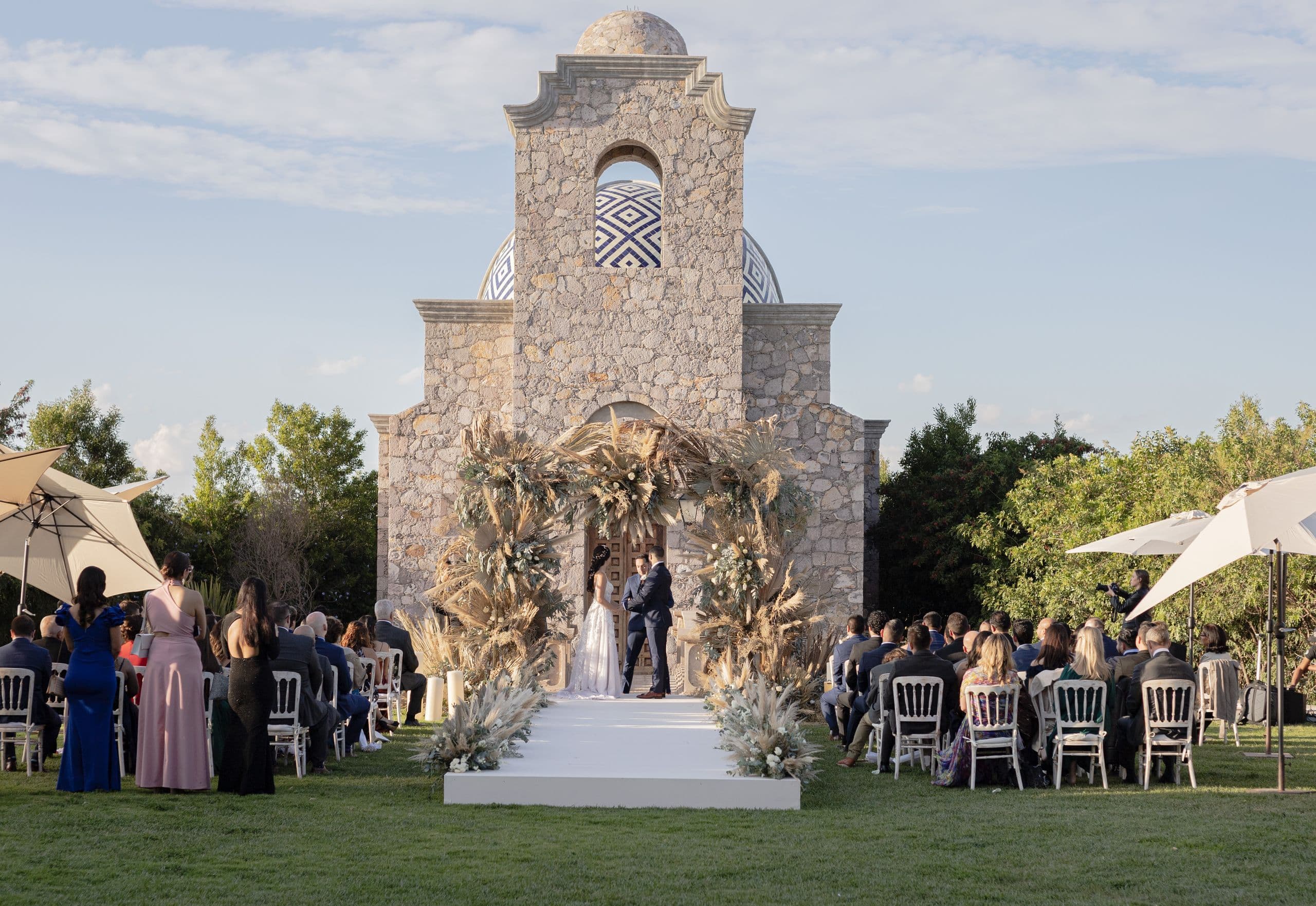 Wedding ceremony with guests and a floral arch in front of a stone hacienda chapel