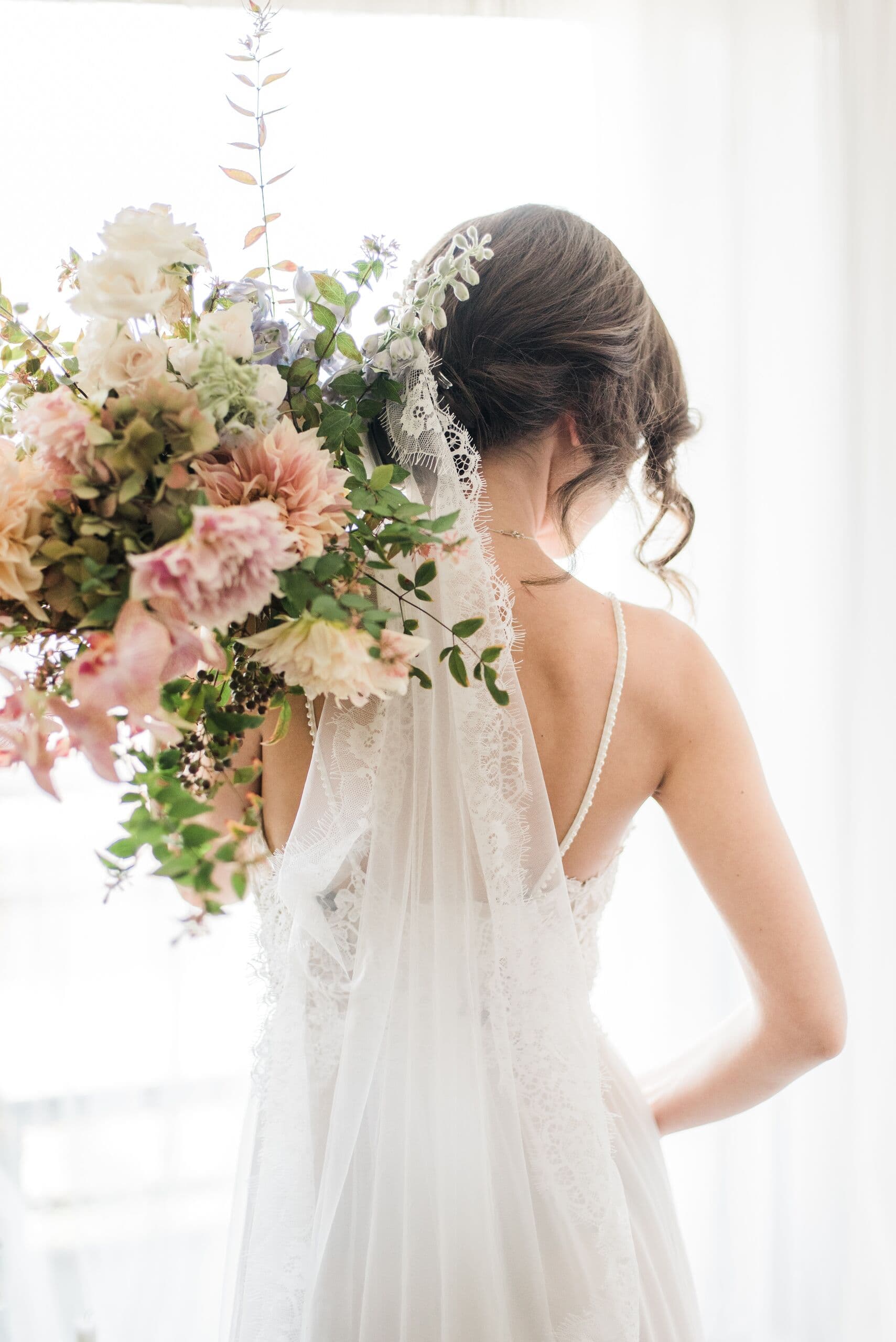 Novia de espaldas con velo, sosteniendo al hombro un ramo en tonos rosa y durazno, a contraluz