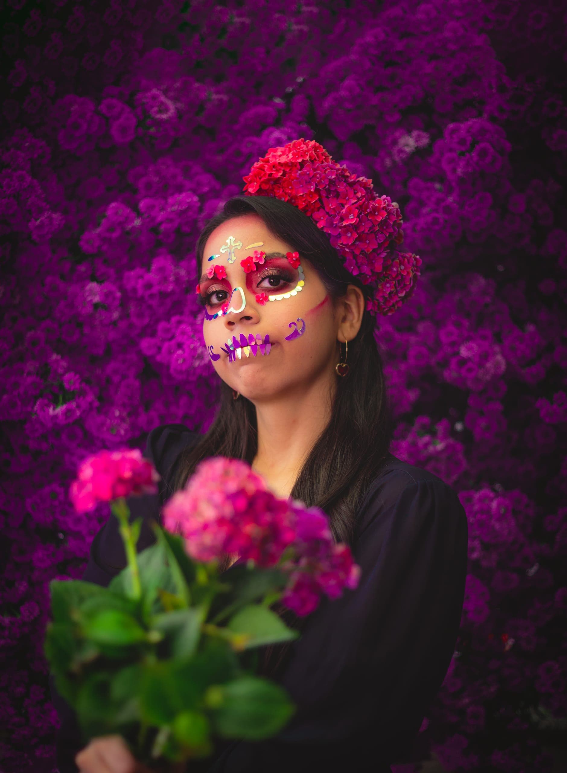 Catrina con corona de hortensias y ramo en mano frente al muro de flores moradas