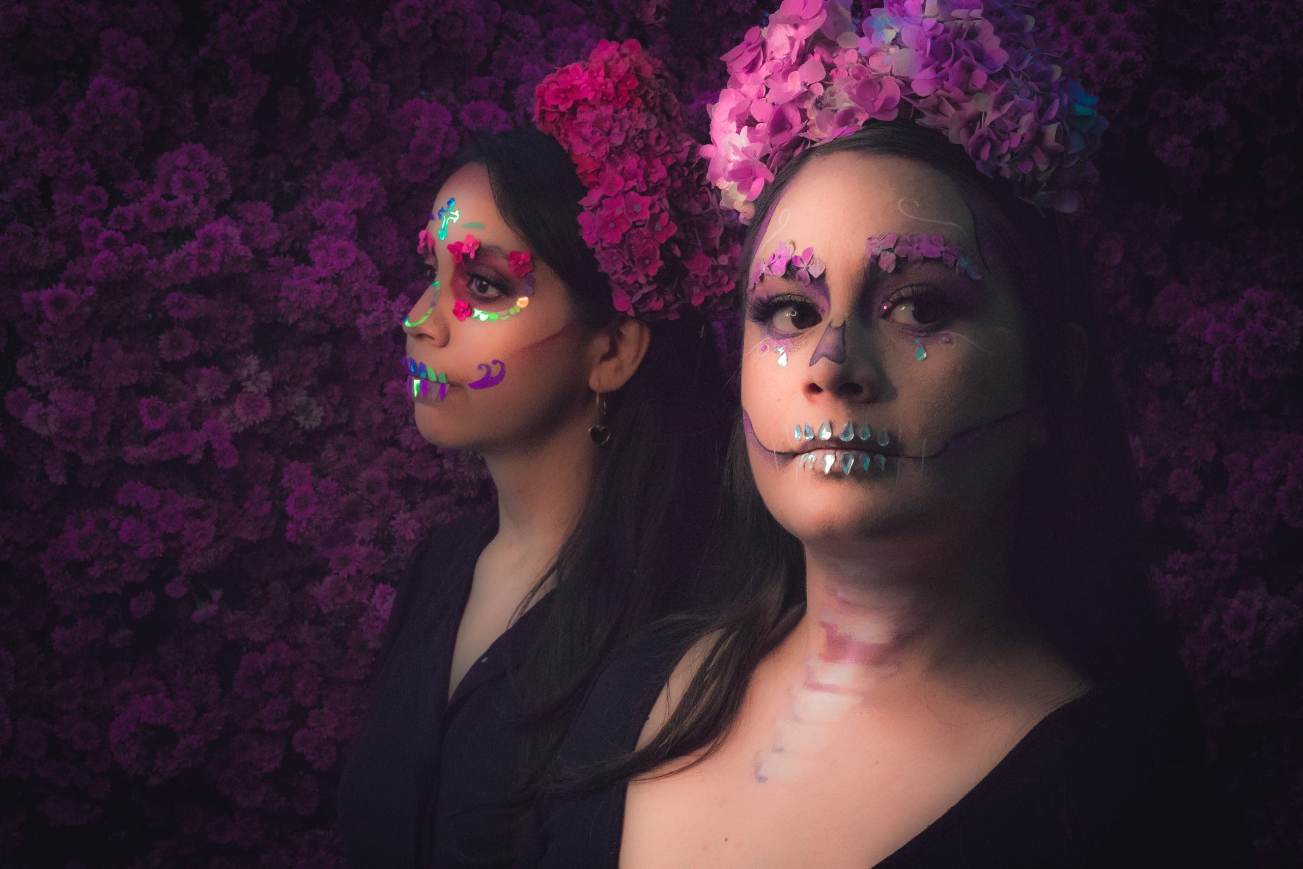 Dos catrinas con coronas florales de perfil frente a un muro de flores