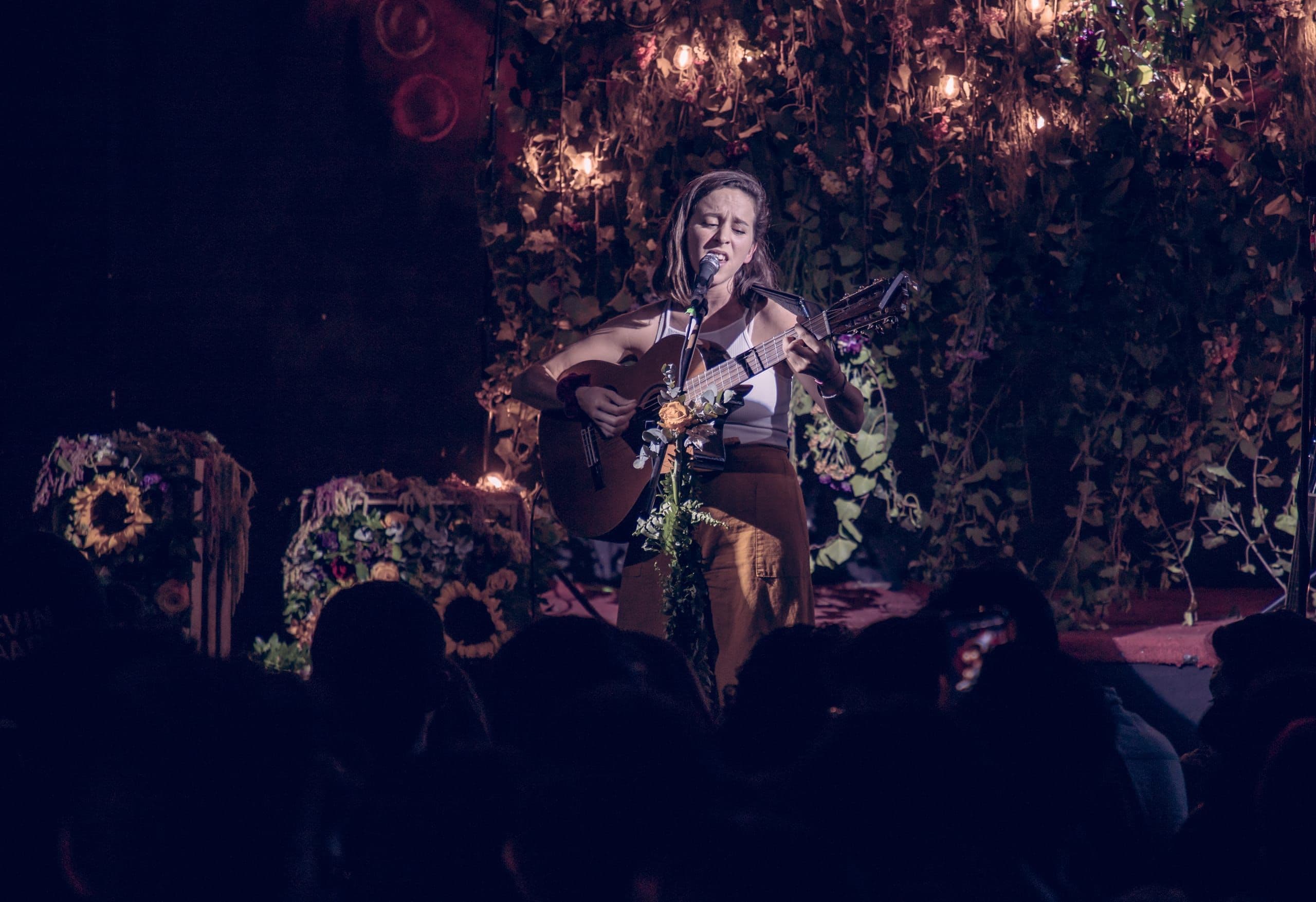 Una artista canta sobre un escenario cubierto de flores y follaje iluminado, ambientado por Clhei