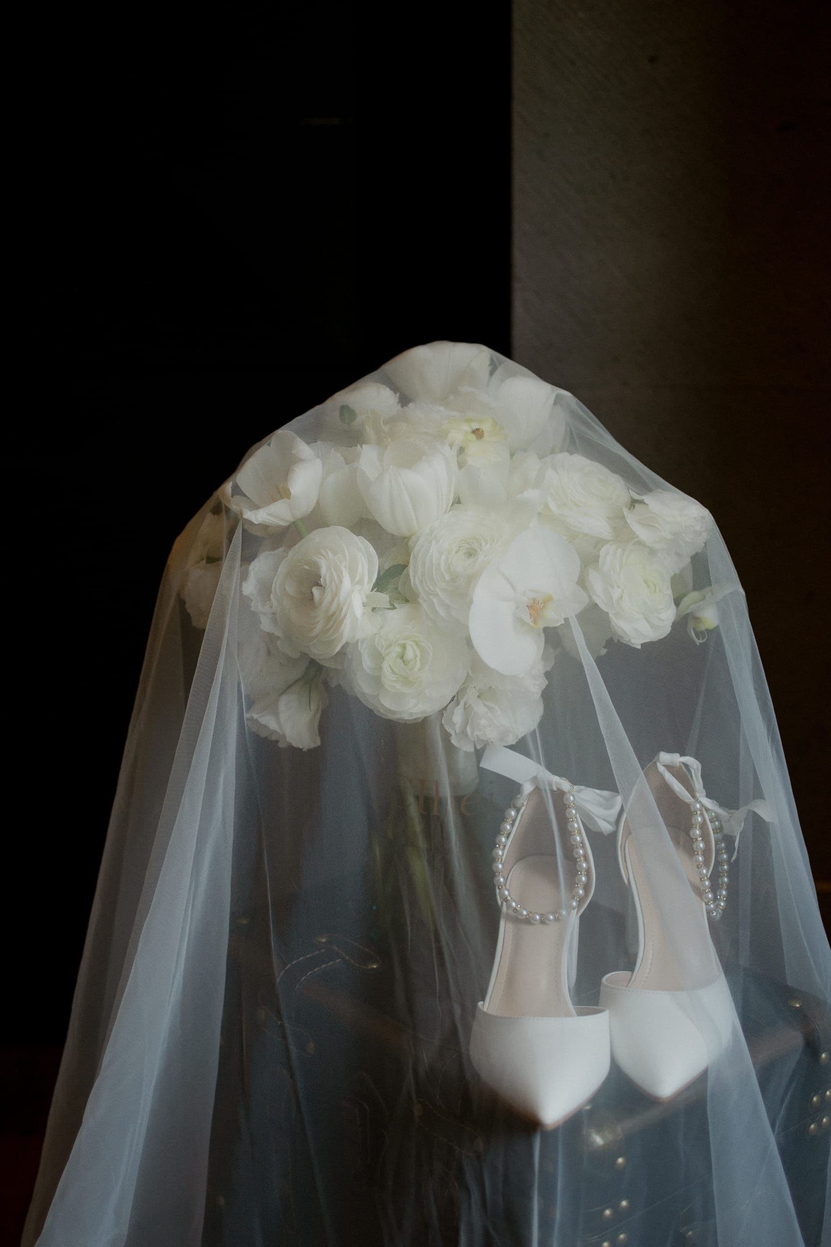 Ramo de novia blanco bajo un velo, junto a zapatos y joyería, en luz de penumbra