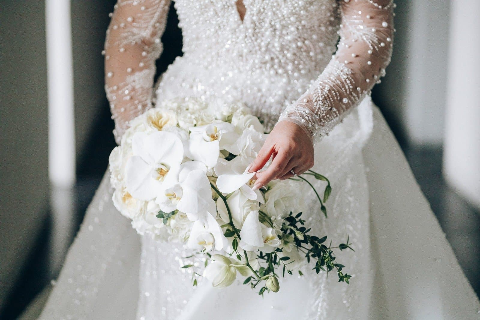 Ramo de orquídeas blancas y follaje fino en manos de una novia con mangas de perlas