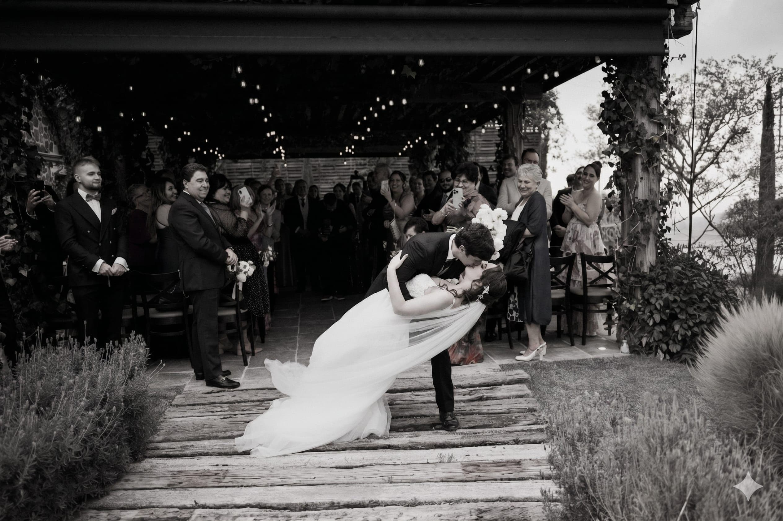 Los novios se besan al final del pasillo mientras los invitados aplauden bajo la pérgola, en blanco y negro