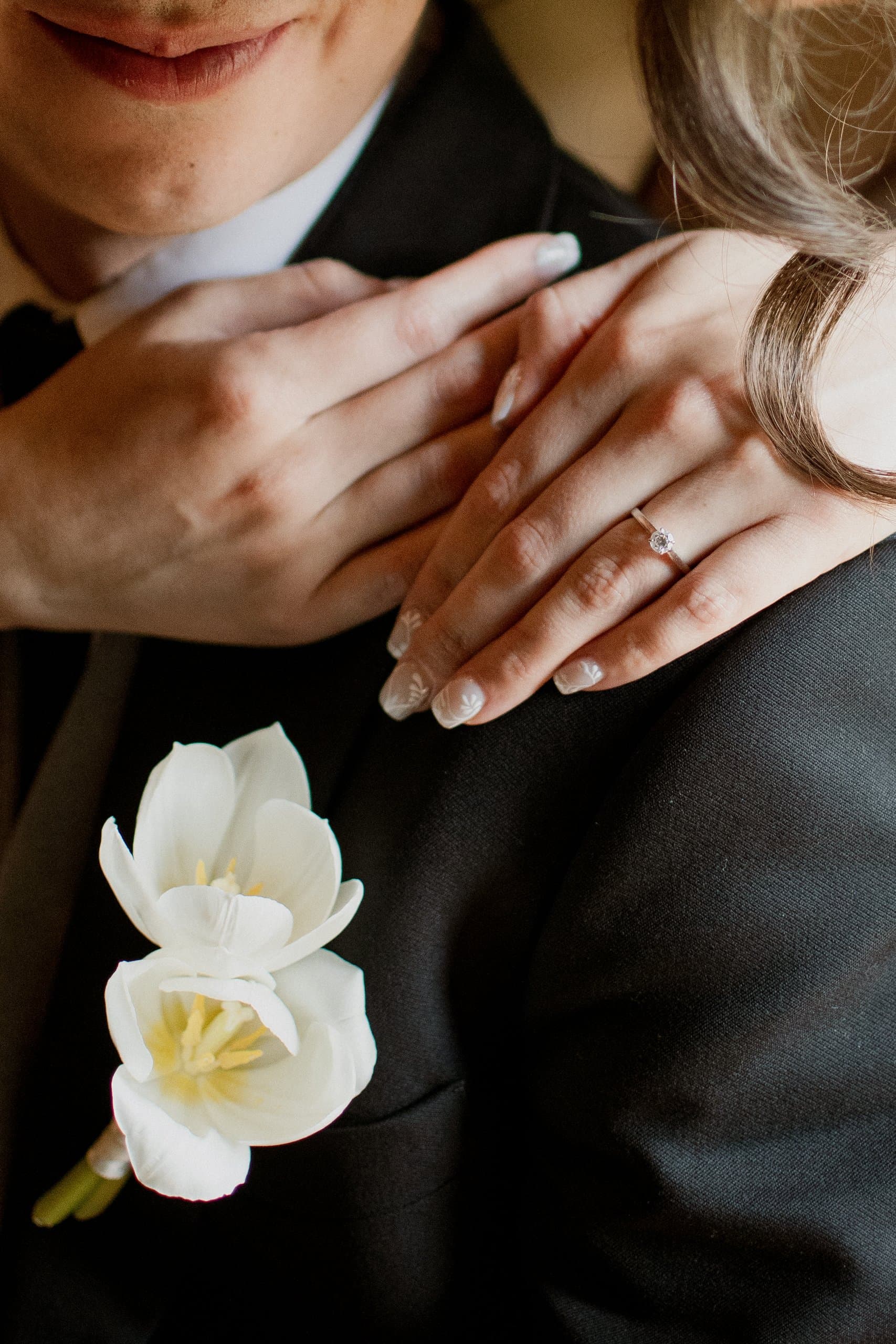 Manos de la pareja sobre el saco del novio, con el anillo y un boutonnière de tulipán blanco