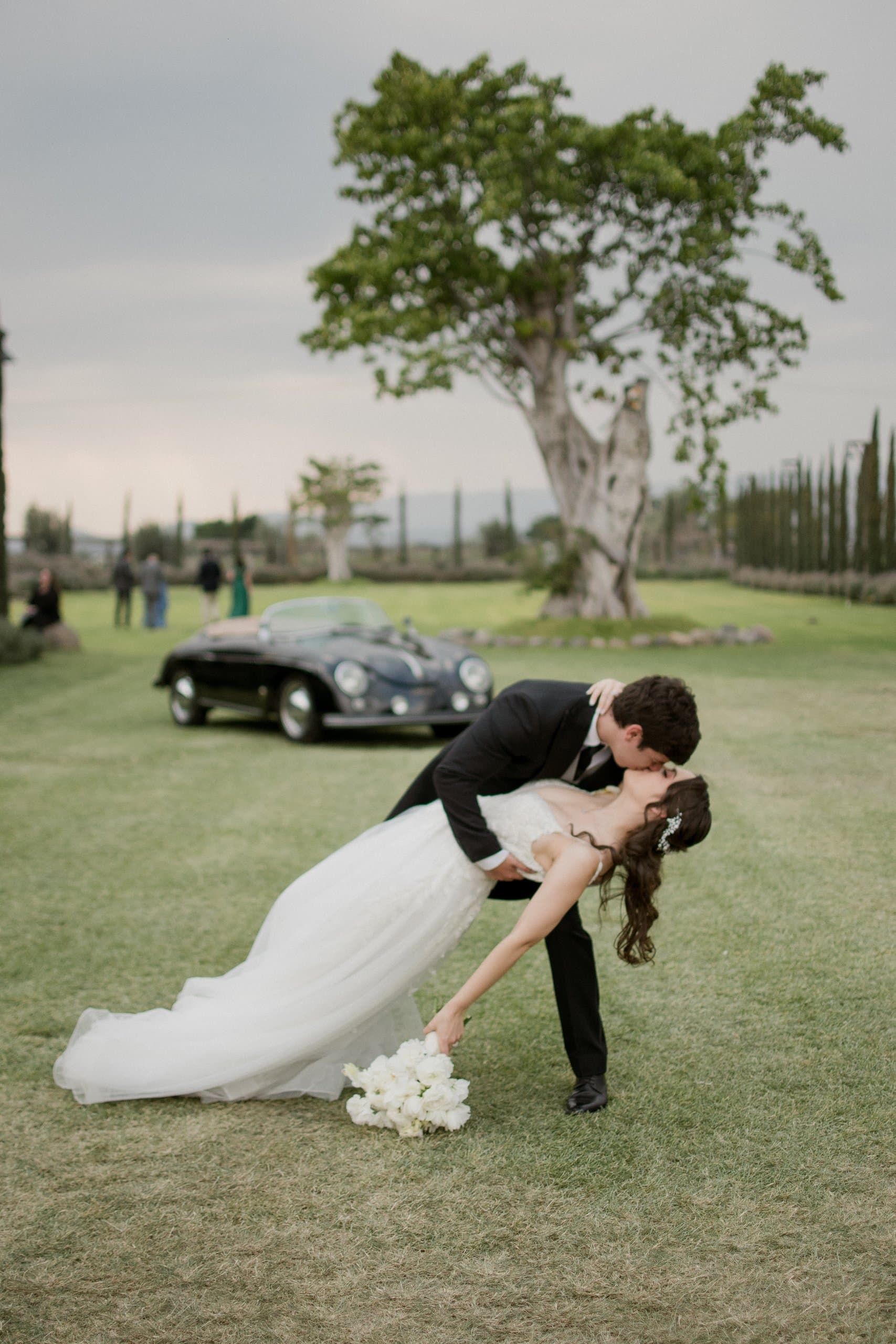 Pareja bailando frente a un árbol y un auto clásico en el jardín, al atardecer