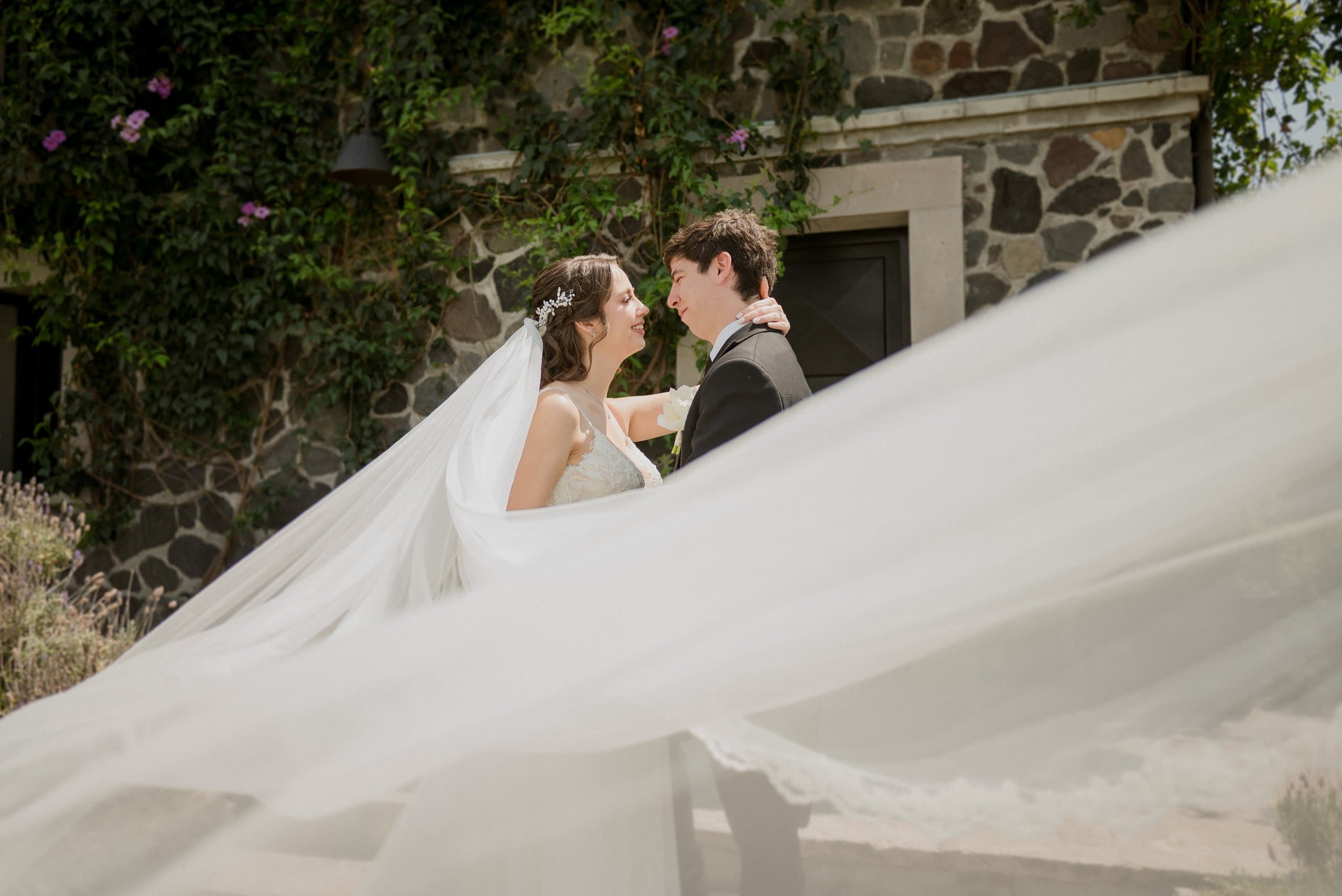 Pareja frente a frente con el velo flotando al viento, junto a un muro de piedra con buganvilias, en una boda planeada por Clhei