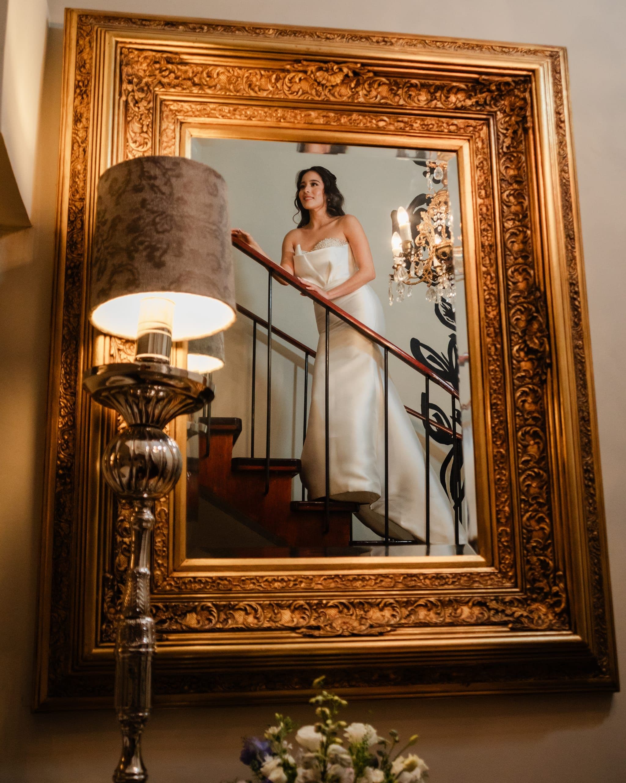 Bride reflected in a gold-framed mirror on the staircase of a Puebla mansion, chandelier above
