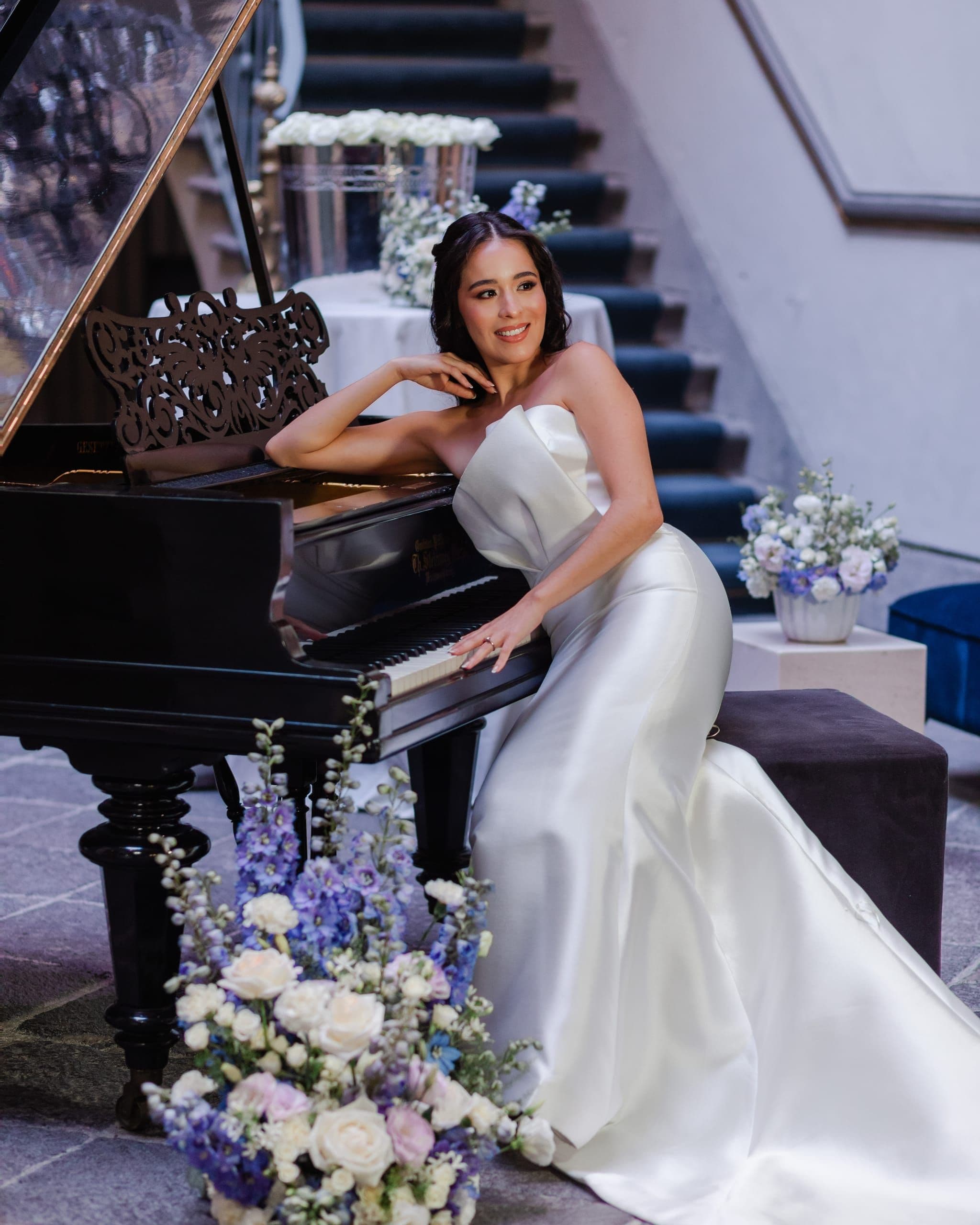 Bride leaning on a grand piano with a white and lavender floral arrangement at her feet